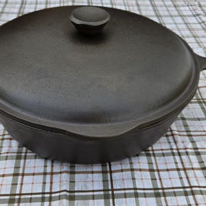 May include: A round, dark gray cast iron skillet with a matching lid and a single handle. The lid features a central knob for lifting, and the skillet has a smooth, deep basin design. The surface of the cookware is matte and uniform in color.