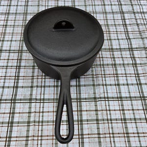 May include: A black cast iron saucepan with a matching lid. The pan features a long, slender handle with a teardrop-shaped cutout at the end. The lid has a small, integrated handle loop in the center. The surface of the cookware is matte black.