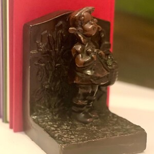 May include: A dark brown metal bookend featuring a sculpted figure of a child standing in front of a textured background with foliage. The bookend supports a small row of books with red, green, and purple spines, providing structural stability.