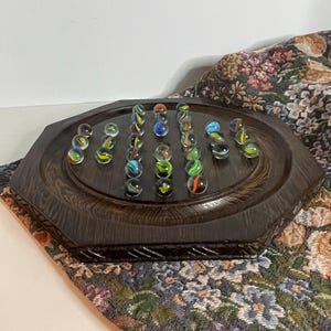 May include: A dark brown wooden octagonal board game featuring a series of small circular indentations arranged in a grid pattern. Several clear glass marbles with colorful internal swirls are placed within the indentations on the board surface.