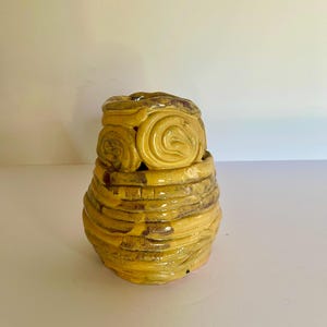 May include: A hand-built ceramic vessel with a glossy yellow glaze and dark brown accents. The base features horizontal coiled layers, while the upper section is decorated with prominent circular spiral motifs. The surface has a textured, uneven finish.