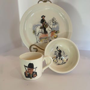May include: A cream-colored ceramic dinnerware set consisting of a plate, a bowl, and a mug. The plate features a cowboy on a white horse. The bowl displays a rider on a horse. The mug shows a portrait of a cowboy holding two revolvers.