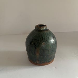 May include: A small, bulbous ceramic vase with a narrow neck and a wide, rounded body. The surface features a mottled, dark teal and forest green glaze with a glossy finish, transitioning to an exposed, unglazed reddish-brown clay base.