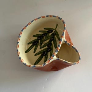 May include: A ceramic dual-compartment serving dish with a cream-colored interior featuring a hand-painted green olive branch with black olives. The rim is decorated with a repeating pattern of small blue and orange squares, and the exterior is terracotta.