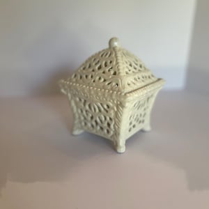 May include: A cream-colored ceramic box with a removable lid, featuring an intricate openwork filigree pattern throughout the body and lid. The square-shaped container stands on four small feet and has a small knob handle on the top center.