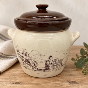 May include: A cream-colored ceramic crock with a dark brown lid and two side handles. The exterior features a brown line-art illustration of a person driving a horse-drawn sleigh through a winter landscape with trees and rolling hills.