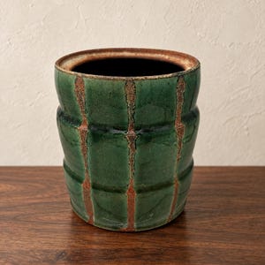 May include: A cylindrical ceramic vessel featuring a deep green glaze with vertical, textured brown stripes. The rim is finished in a contrasting brown tone, and the body has subtle horizontal indentations along its exterior surface.