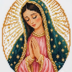 Pode incluir: Um bordado em ponto cruz representando a Virgem de Guadalupe com as mãos juntas em oração. Ela veste uma túnica rosa e um manto azul-petróleo adornado com estrelas douradas, cercada por um padrão de raios de sol dourados em fundo branco.