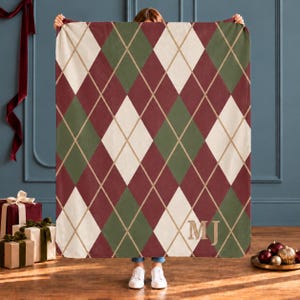 May include: A rectangular fleece blanket featuring a repeating argyle pattern in shades of deep red, olive green, and cream, accented by thin gold lines. The bottom right corner displays the embroidered gold monogram letters MJ.