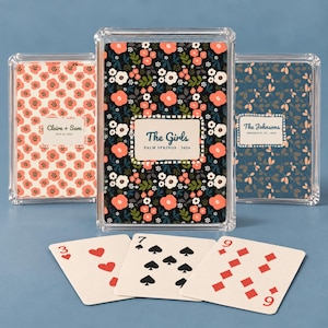 May include: Three clear plastic cases containing custom playing cards with floral patterns. The center case features a black background with coral and white flowers and the text The Girls PALM SPRINGS 2024. Three loose cards are displayed in front.