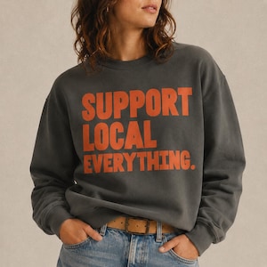 May include: A charcoal grey crewneck sweatshirt featuring the text SUPPORT LOCAL EVERYTHING. printed in bold orange capital letters across the chest. The garment has long sleeves and a ribbed neckline, cuffs, and hem, providing a classic fit.
