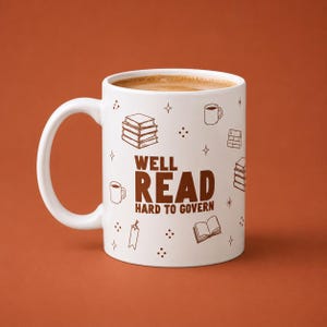 May include: A white ceramic mug featuring brown line art illustrations of books, mugs, and stars. The text "WELL READ HARD TO GOVERN" is printed in large brown letters in the center. The mug is filled with a light brown liquid, likely coffee or tea.