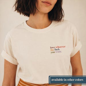 May include: A cream-colored short-sleeve t-shirt with a crew neckline and rolled sleeves. The chest features the text "love whoever the fuck you want." printed in a multi-colored serif font with red, orange, blue, and purple lettering.