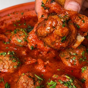 May include: A serving of round, brown meatballs simmered in a thick, vibrant red tomato sauce, garnished with finely chopped green herbs. One meatball is lifted out of the bowl on a piece of crusty white bread, showing the textured surface.