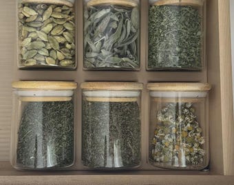 Puede incluir: Seis frascos de vidrio transparente con tapas de bambú claro dispuestos en un cajón. Los frascos contienen diversas hierbas y especias secas, incluyendo vainas de cardamomo verde, hojas de salvia y pequeñas flores de manzanilla blancas.