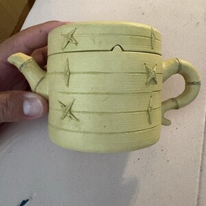 May include: A pale yellow ceramic teapot with a cylindrical body and a lid. The exterior features horizontal grooved lines and small, raised star-shaped decorative accents. It includes a curved spout and a handle shaped like bamboo segments.