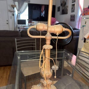 May include: A metallic table lamp base with a weathered gold finish, featuring a central column, a decorative pedestal base, and two light bulb sockets. The lamp includes two pull chains for operation and an exposed electrical cord.