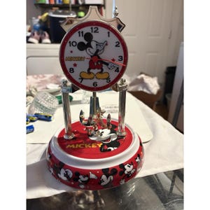 May include: A Mickey Mouse themed analog clock with a circular white face, red border, and black numerals. The clock features a silver-toned frame, a rotating pendulum, and a red base decorated with multiple Mickey Mouse illustrations and the name Mickey.