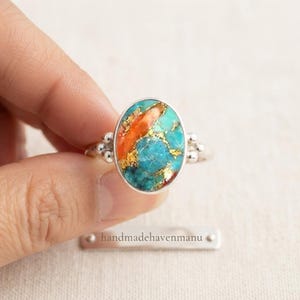Könnte beinhalten: Ein Silberring mit einem ovalen Stein in einem lebendigen türkisfarbenen und orangefarbenen Marmormuster, akzentuiert durch goldfarbene Sprenkel. Die Ringschiene hat ein Perlendesign auf jeder Seite der Fassung. Der Text handmadehavenmanu ist zu sehen.