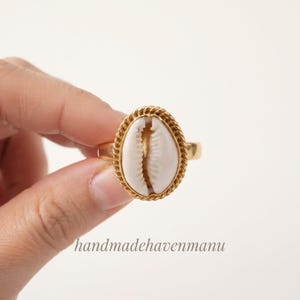 Peut inclure: Une bague dorée ornée d'un coquillage cauri blanc central serti dans un bord en métal torsadé. La bague possède un anneau doré lisse et poli, présentée sur un fond uni avec le texte handmadehavenmanu inscrit en bas de l'image.