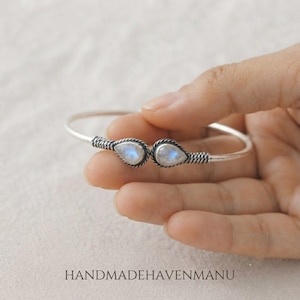 May include: A silver cuff bracelet featuring two teardrop-shaped moonstones at the open ends. The metal band has a twisted rope texture, and the stones display a soft blue iridescent glow. The text HANDMADEHAVENMANU is visible.