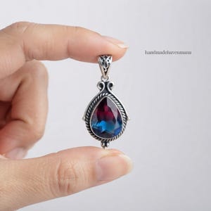 May include: A teardrop-shaped pendant featuring a faceted gemstone with a gradient transition from deep magenta to blue. The stone is encased in a silver setting with a twisted rope border and an ornate, filigree-style bail at the top.