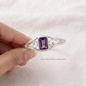May include: A silver-toned cuff bracelet featuring a central emerald-cut purple gemstone. The metal band displays an openwork design with swirling scroll patterns on both sides of the stone. The text handmadehavenmanu is visible.