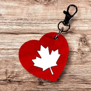 May include: A heart-shaped keyring featuring a vibrant red background with a prominent white maple leaf design in the centre. The wooden heart is attached to a black metal lobster clasp by a small circular metal ring, providing a secure attachment.
