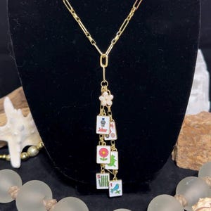 May include: A gold-toned chain necklace featuring a pendant arrangement of small, rectangular mahjong tiles. The tiles display various colorful illustrations, including a flower, a bird, and a joker, suspended from a small white flower charm.