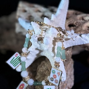 May include: A gold-toned bracelet featuring jade-colored beads and charms with Mahjong tile designs. The bracelet includes white flower and key charms, with a toggle clasp. The background is dark, highlighting the jewelry.