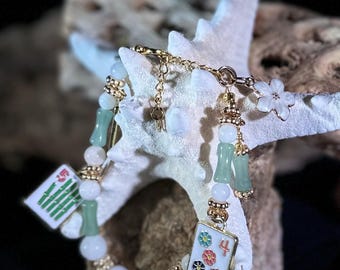 Elegante bracciale con ciondoli a forma di tessere di Mahjong, finiture placcate in oro 18 carati, pietra color giada verde, perline di conchiglia e divertenti dettagli in smalto.