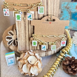 May include: Two gold-toned chain link bracelets featuring dangling mahjong tile charms. The charms display colorful illustrations of flowers, a blue bird, green bamboo stalks, and Chinese characters. Each bracelet is displayed on a brown card.