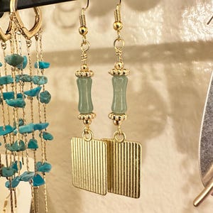 Pu&ograve; includere: Un paio di orecchini pendenti color oro con una perla cilindrica verticale verde chiaro accentuata da piccoli tappi in metallo dorato, sospesi sopra un ciondolo rettangolare dorato con una sottile trama a coste verticali.