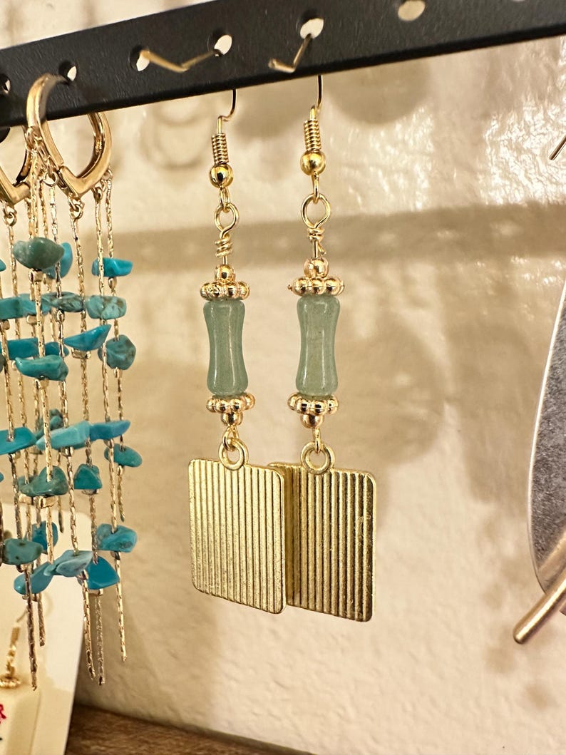 Pu&ograve; includere: Un paio di orecchini pendenti color oro con una perla cilindrica verticale verde chiaro accentuata da piccoli tappi in metallo dorato, sospesi sopra un ciondolo rettangolare dorato con una sottile trama a coste verticali.