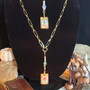 May include: A gold-toned chain necklace featuring two rectangular pendants resembling mahjong tiles. The top pendant displays a blue and white design, while the bottom pendant features a red flower. Each tile is accented with decorative beads.