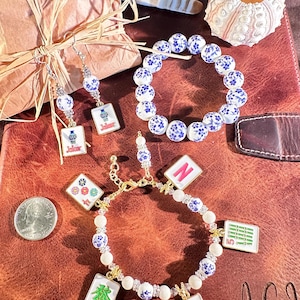 May include: A jewelry set featuring a beaded bracelet and matching earrings with blue and white floral patterns, plus a charm bracelet with mahjong tile pendants including a pink N, a green 5, a red flower, and a multi-colored floral tile.