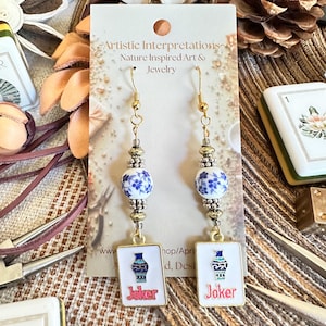 May include: A pair of dangle earrings featuring gold-tone hooks, round white beads with blue floral patterns, silver-tone spacers, and rectangular white charms. Each charm displays a blue and green vase illustration above the red text Joker.