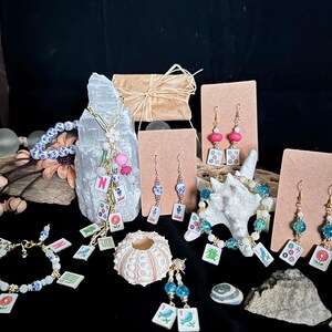 Pu&ograve; includere: Una collezione di gioielli con tessere del mahjong in miniatura, che include collane, bracciali e orecchini. I pezzi incorporano perline colorate, motivi floreali e componenti dorati, esposti accanto a elementi naturali come cristalli e conchiglie.