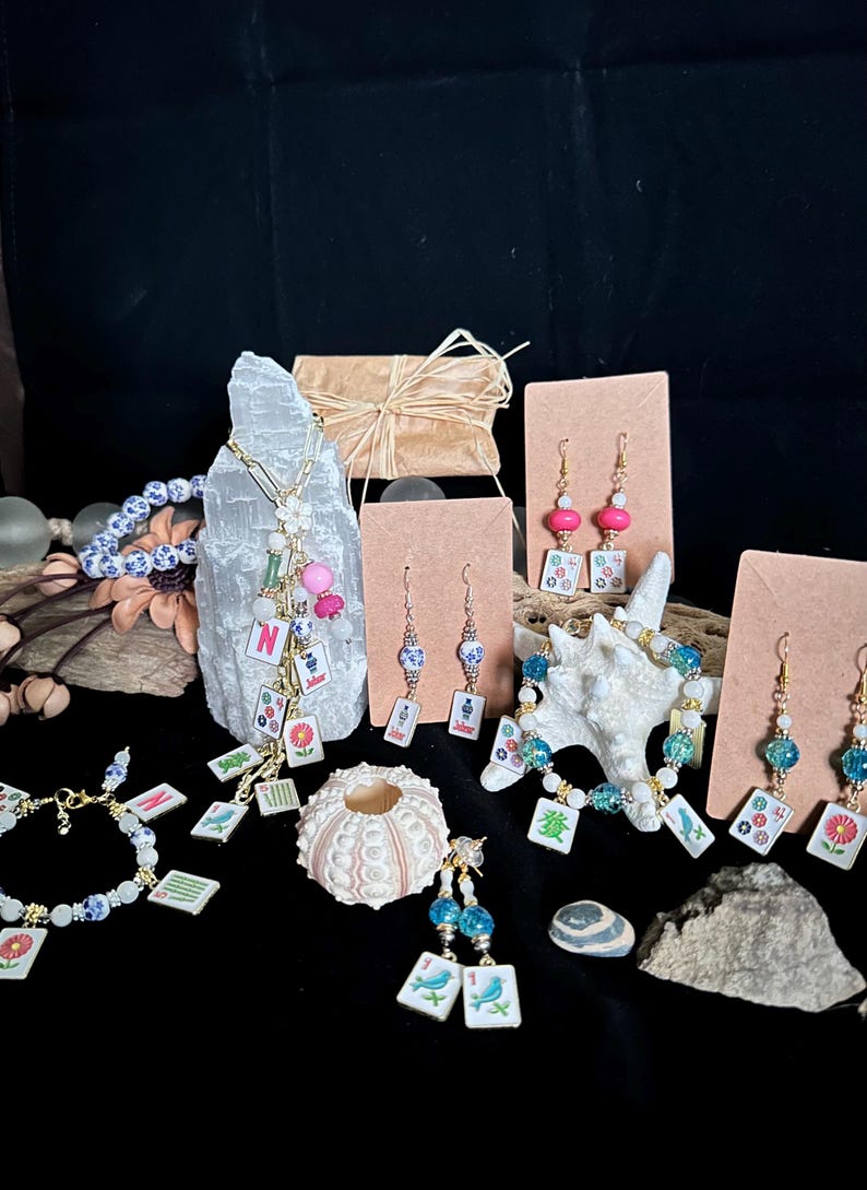 Pu&ograve; includere: Una collezione di gioielli con tessere del mahjong in miniatura, che include collane, bracciali e orecchini. I pezzi incorporano perline colorate, motivi floreali e componenti dorati, esposti accanto a elementi naturali come cristalli e conchiglie.