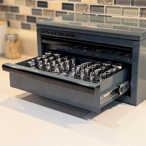 Peut inclure: Une boîte à outils en métal gris de marque U.S. General avec un tiroir ouvert. Le tiroir contient une collection d'embouts de clé à douille en métal argenté disposés en rangées. La boîte présente une finition lisse et brillante et une poignée noire.