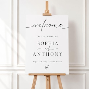 Può includere: Un cartello di benvenuto per matrimonio rettangolare bianco su un cavalletto in legno. Il cartello presenta un testo nero che recita: welcome TO OUR WEDDING SOPHIA and ANTHONY August 17th, 2024 | Galena, Illinois, con un piccolo cuore disegnato.