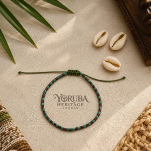 May include: A bracelet featuring alternating dark red and teal cylindrical beads on a thin olive green cord with an adjustable sliding knot closure. The bracelet is positioned near three white cowrie shells and the text Yoruba Heritage Studio.
