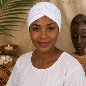 May include: A solid white turban headwrap featuring a gathered, knotted front design. The fabric is smooth and opaque, covering the crown of the head completely. The text YORUBA HERITAGE STUDIO is visible in the upper right corner of the image.