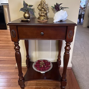 May include: A dark brown wooden side table with a single drawer, fluted legs, and oval decorative accents. The top surface holds a brass Ganesha statue, a small brass vase with green leaves, and a white conch shell. A silver bowl with red petals sits on the lower shelf.
