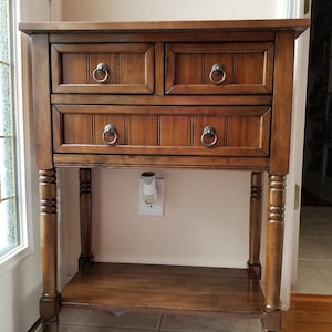 May include: A medium-brown wooden side table featuring two small top drawers and one wider bottom drawer, all equipped with circular metal ring pulls. The table stands on four turned legs and includes a lower shelf for storage.