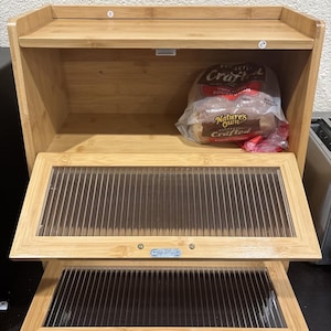 May include: A two-tier bamboo bread box with a natural light wood finish. The unit features two drop-down doors with ribbed, translucent plastic panels and silver-toned metal hardware. A loaf of Nature's Own Crafted sourdough bread sits inside.