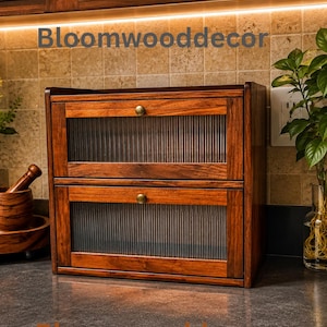 May include: A two-tier wooden bread box with a dark brown finish, featuring two flip-down doors with fluted glass panels and small brass-toned knobs. The text Bloomwooddecor is visible on the front. The unit has a rectangular frame.