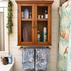 May include: A wooden wall-mounted cabinet with two glass doors and brass handles, featuring interior shelves holding various toiletries. Below the cabinet is a brass towel bar holding two blue towels with a sea turtle and coral pattern.