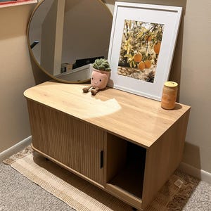 May include: A light wood cabinet with a sliding tambour door and an open side compartment. On top sits a round gold-rimmed mirror, a framed print of oranges on a tree, a small plush succulent toy with a smiling face, and a yellow candle jar.
