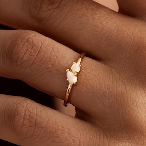 May include: A thin gold ring featuring two small, oval-shaped white opals set side-by-side in a delicate prong setting. The band is smooth and polished, showcasing a minimalist design that highlights the iridescent stones on the finger.
